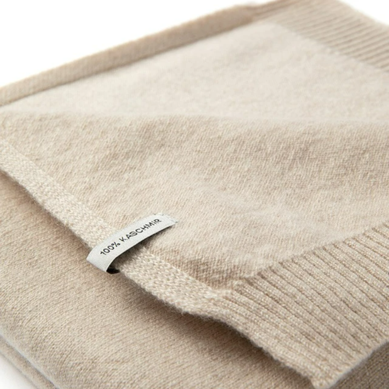 Gefaltete Kaschmirdecke in beige auf weißem Hintergrund, mit sichtbarem Etikett: 100% Kaschmir