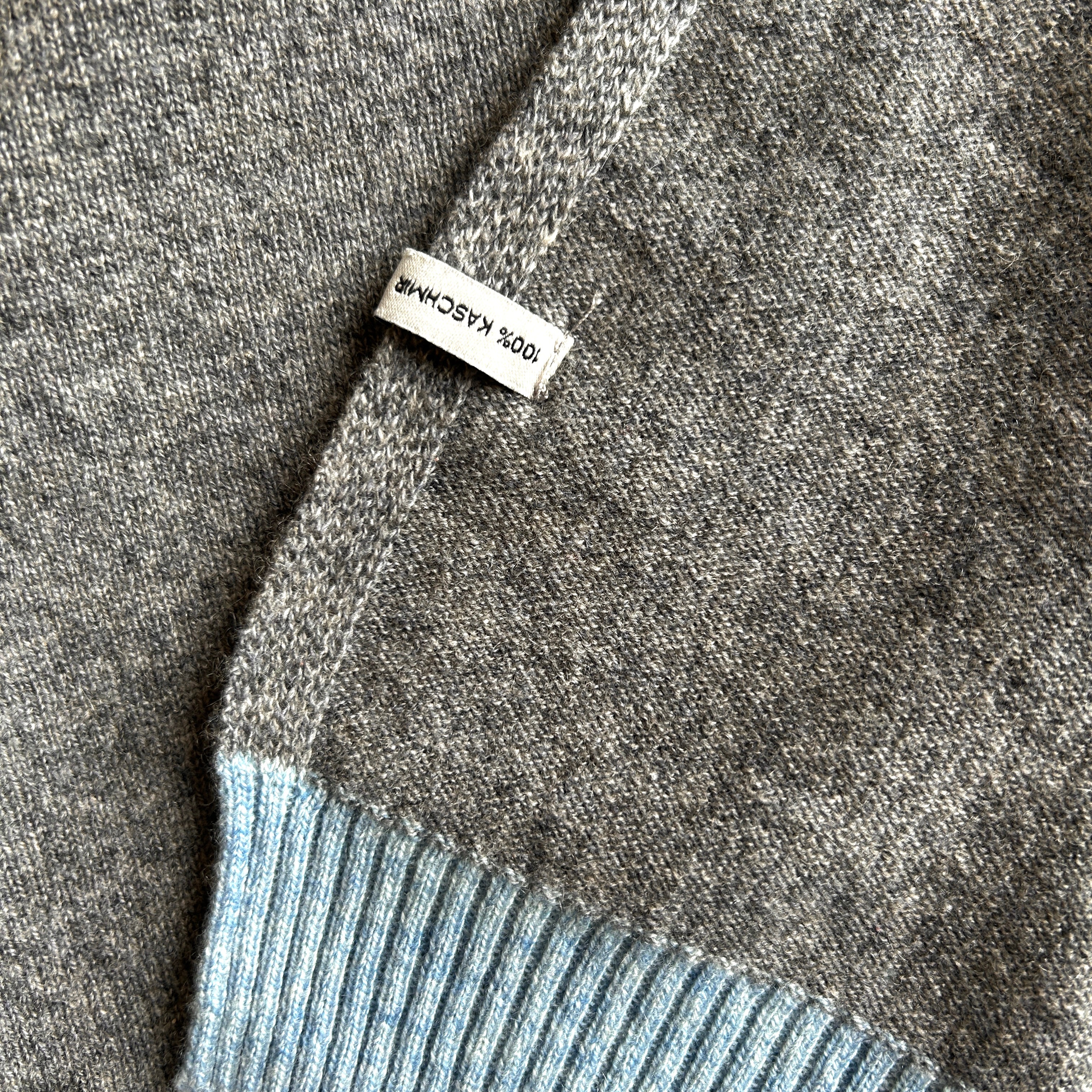 Ausschnitt einer Kaschmirdecke in Grau mit Blauem Rand. Der Etikett mit Namen und Material ist falsch aufgenäht.