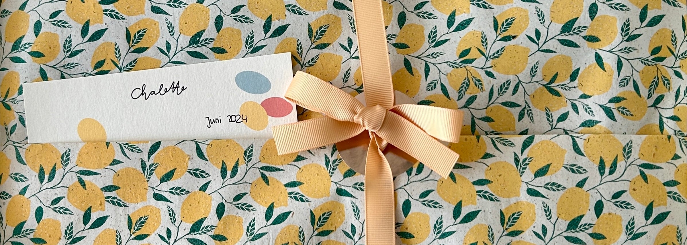 Geschenkverpackung mit Zitronenmusterpapier und beigefarbener Schleife, mit handgeschriebener Karte