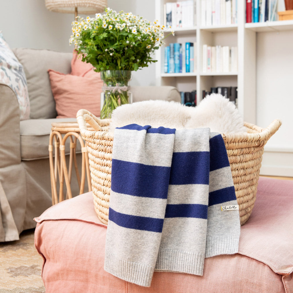 Babydecke in grau mit blauen Streifen über einem Korb mit Lammfell, auf rosafarbenem Pouf im Wohnzimmer drapiert