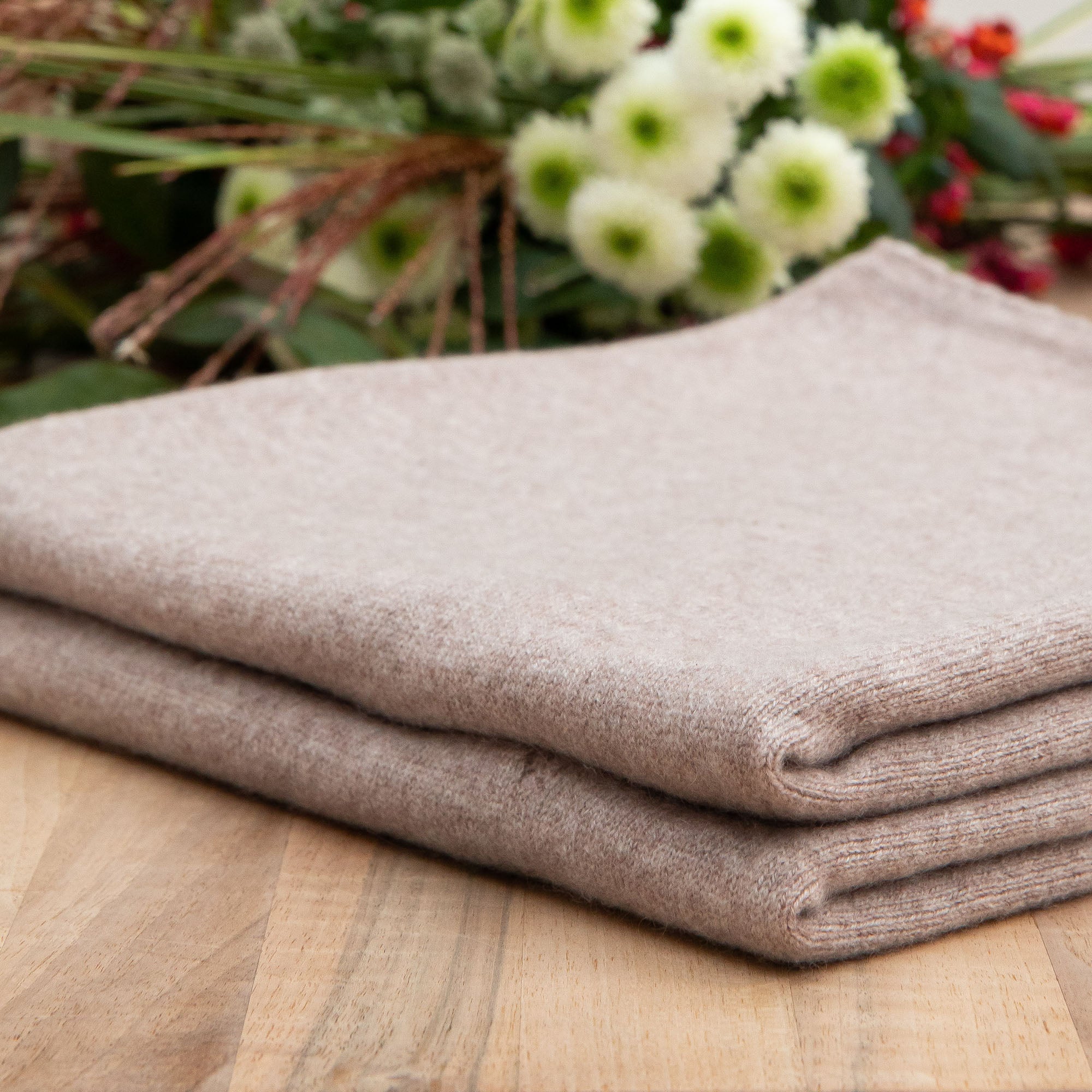 Nahaufnahme einer Kaschmirdecke mit Lochmuster in beige, gefaltet auf einem Holztisch, Hintergrund mit Blumen