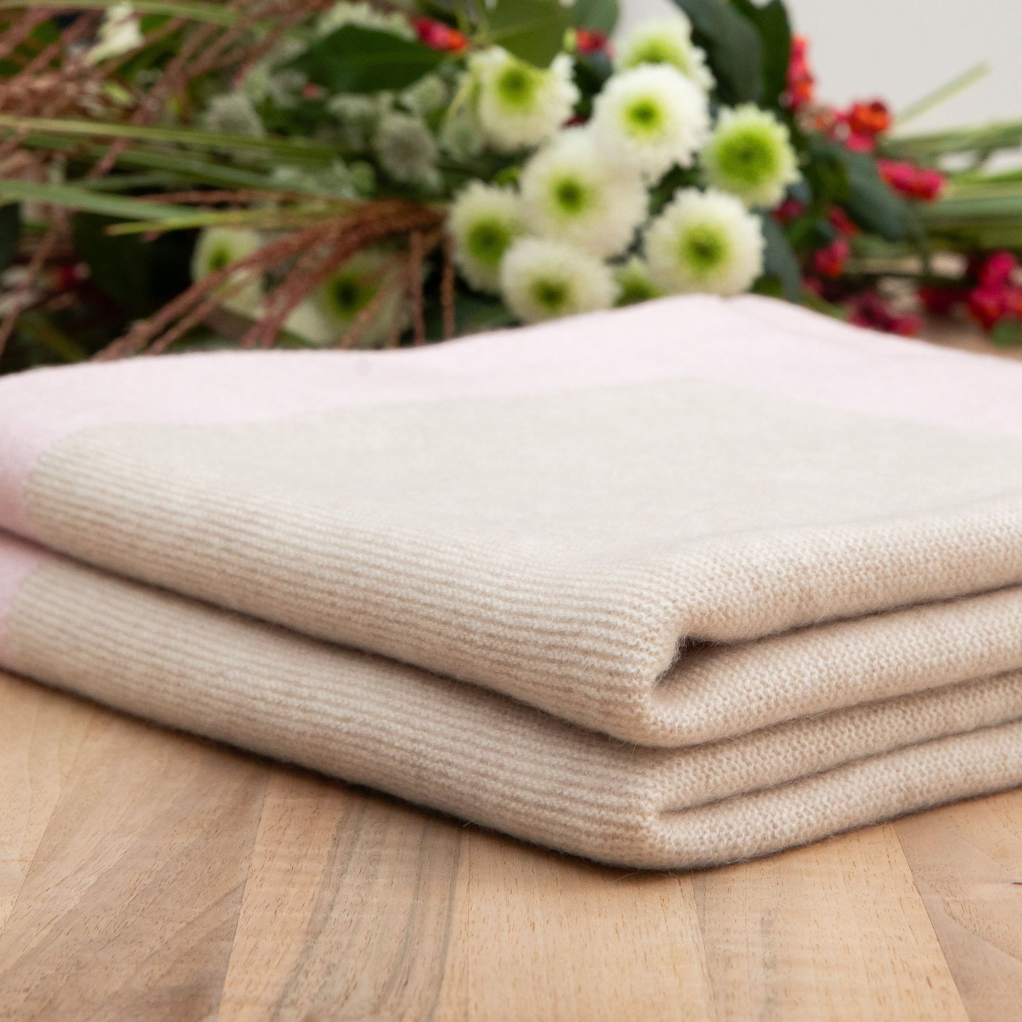 Gefaltete Kaschmirdecke in beige mit rosa Rand mit feinem Strickmuster auf Holztisch, Hintergrund mit Blumen