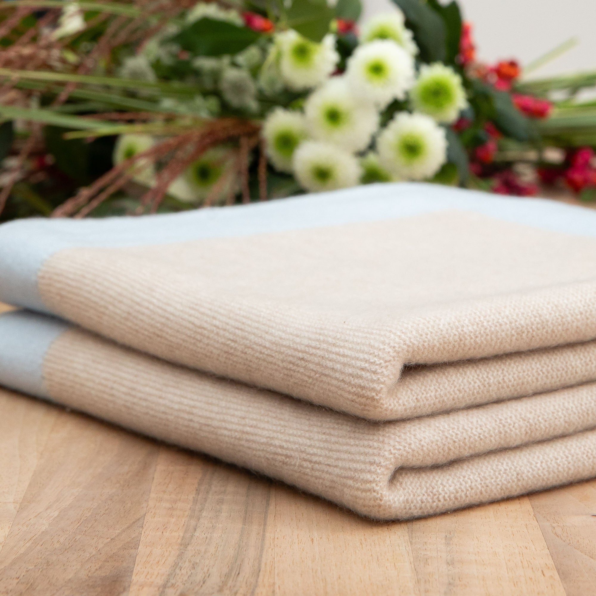 Gefaltete Kaschmirdecke in beige mit blauem Rand mit feinem Strickmuster auf Holztisch, Hintergrund mit Blumen