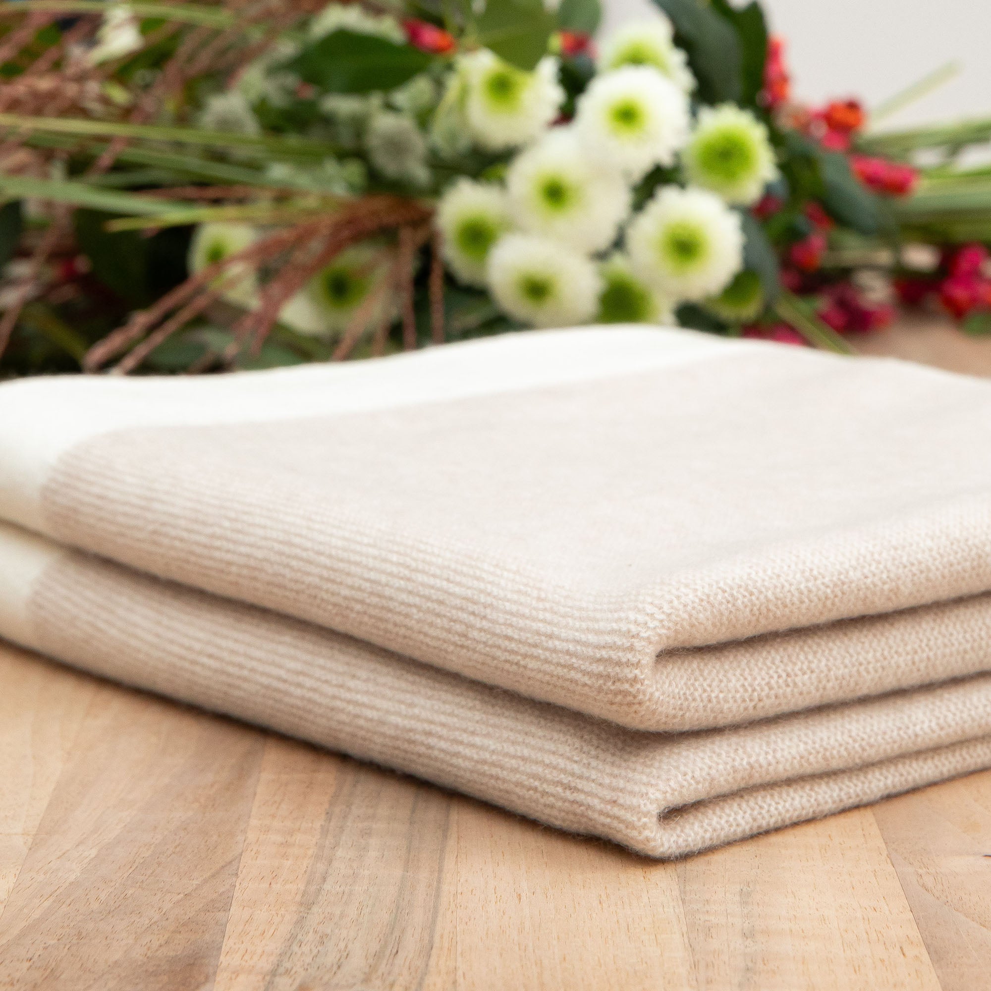 Nahaufnahme einer beigen Kaschmirdecke mit rosa Rand, gefaltet auf einem Holztisch, Hintergrund mit Blumen