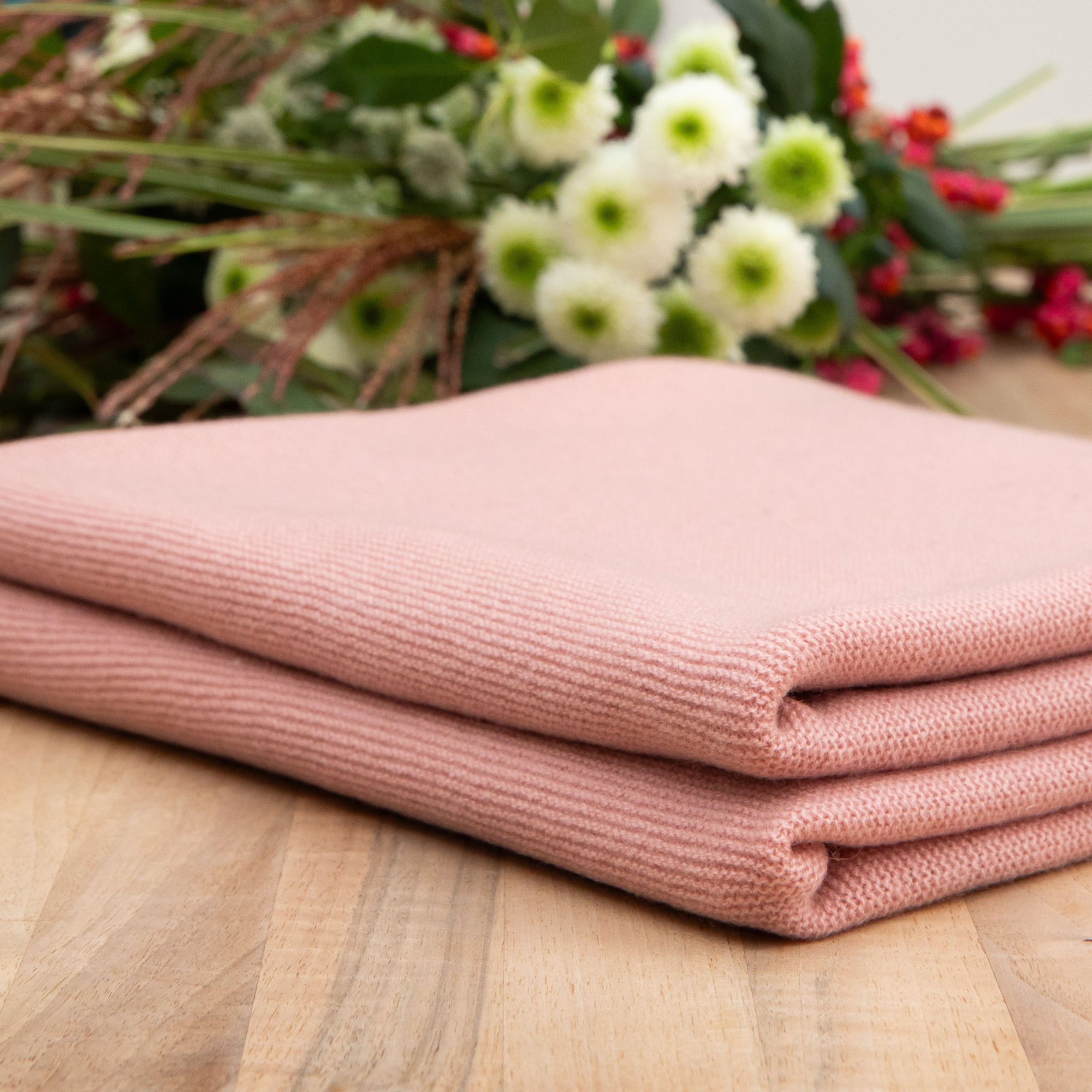 Gefaltete Babydecke in rosa auf Holztisch, Hintergrund mit Blumen