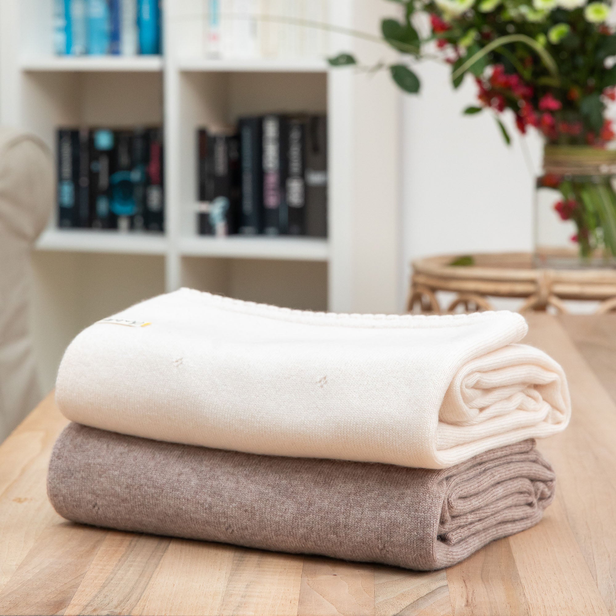 Gefaltete Babydecken aus Kaschmir in off-white und beige mit Lochmuster, Hintergrund mit Bücherregal und Blumen