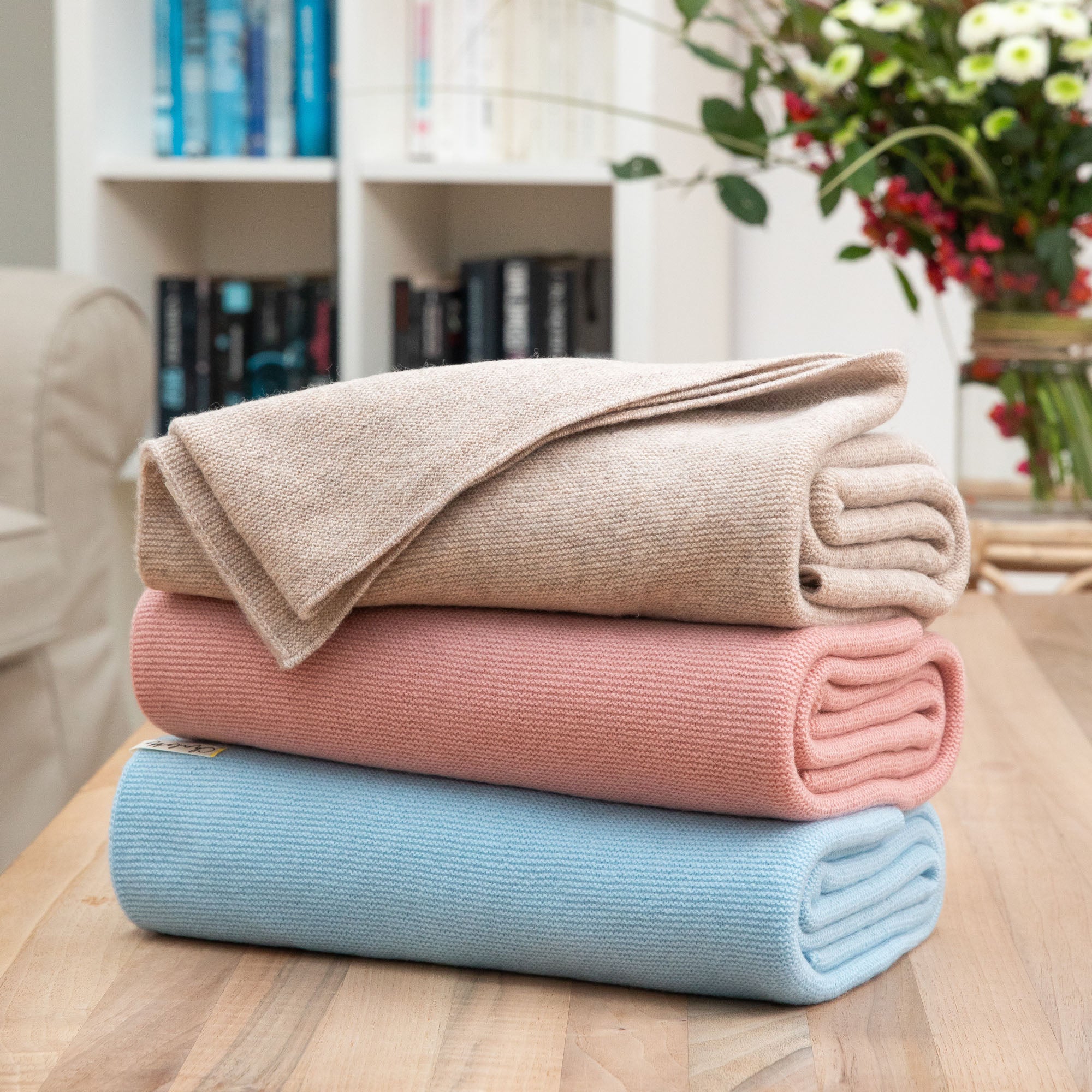 Gefaltete Babydecken in hellblau, rosa und beige liegen übereinander auf einem Holztisch, Hintergrund mit Blumen und Bücherregal