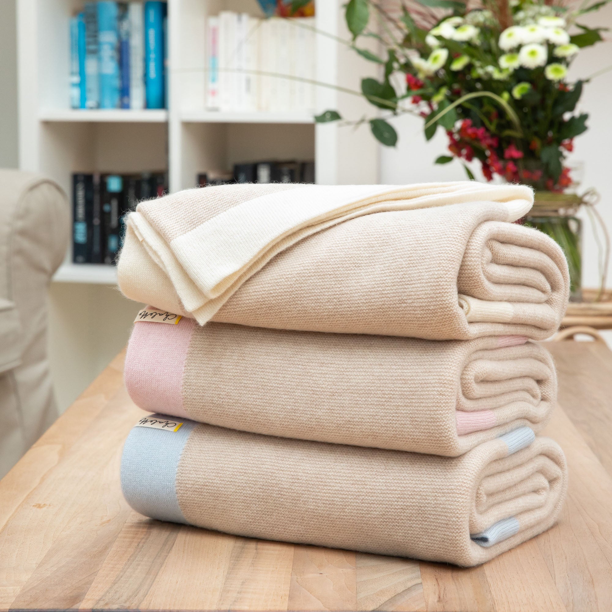 Gefaltete Kaschmirdecken in beige mit blauem, rosa und off-whiten Rand auf Holztisch, Hintergrund mit Bücherregal und Blumen