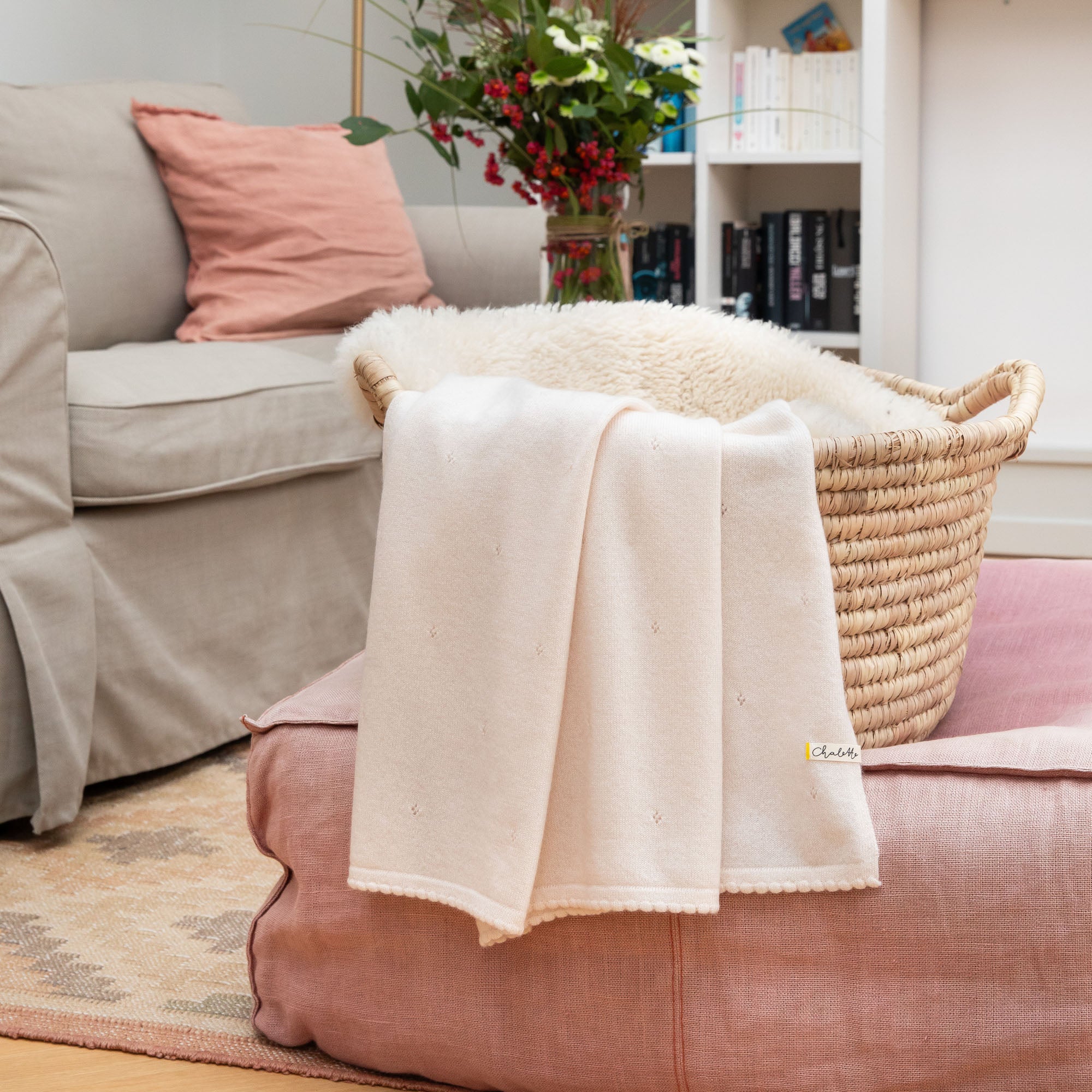 Kaschmirdecke in off-white mit Lochmuster über einem Korb mit Lammfell, auf rosafarbenem Pouf im Wohnzimmer drapiert