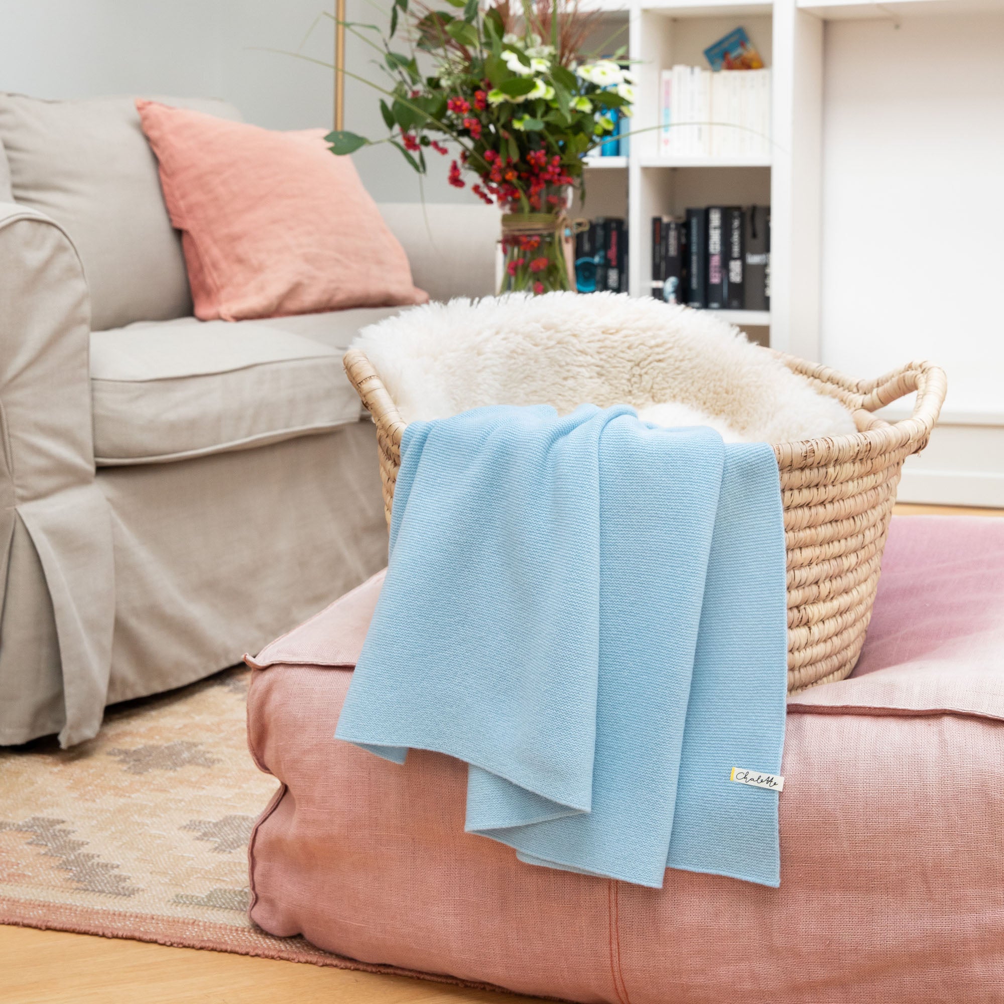 Babydecke aus Kaschmir und Merinowolle in blau über einem Korb mit Lammfell, auf rosafarbenem Pouf im Wohnzimmer drapiert