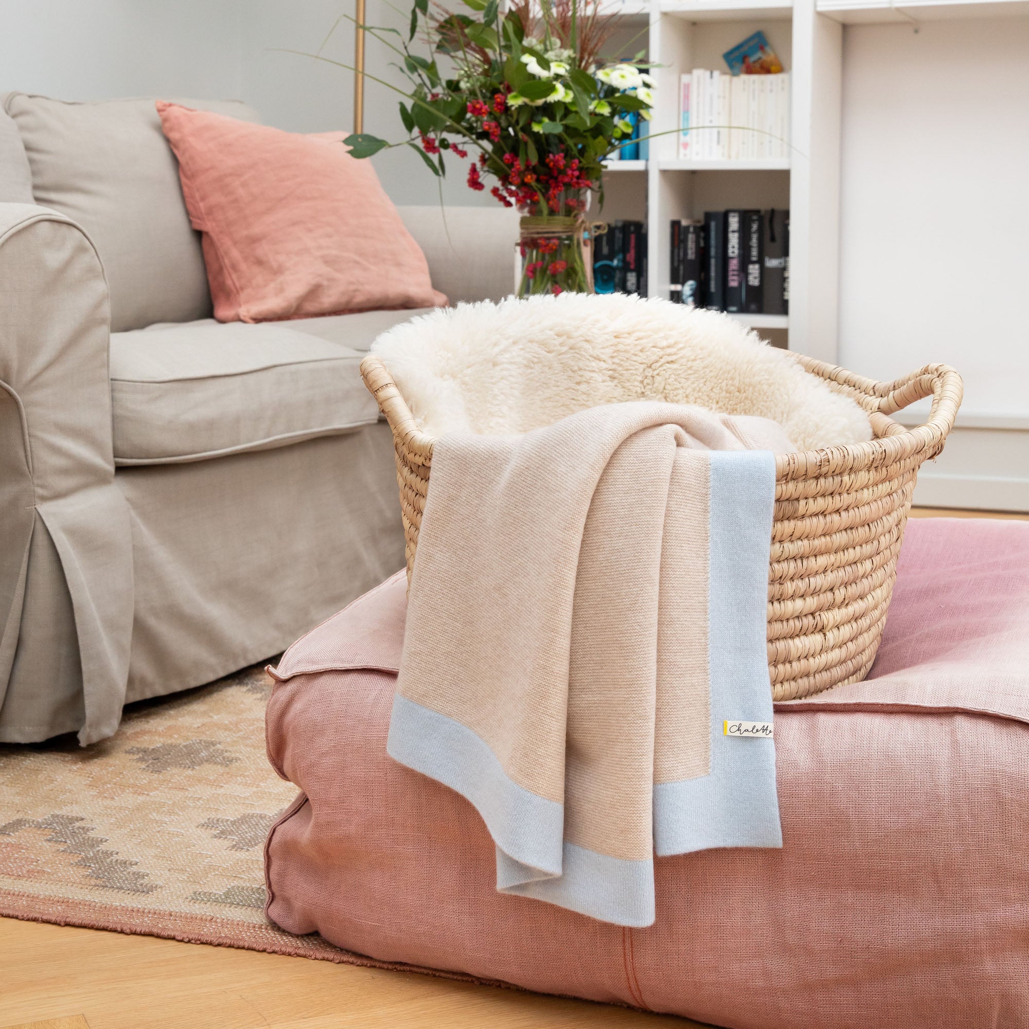 Kaschmirdecke in beige mit blauem Rand über einem Korb mit Lammfell, auf rosafarbenem Pouf im Wohnzimmer drapiert