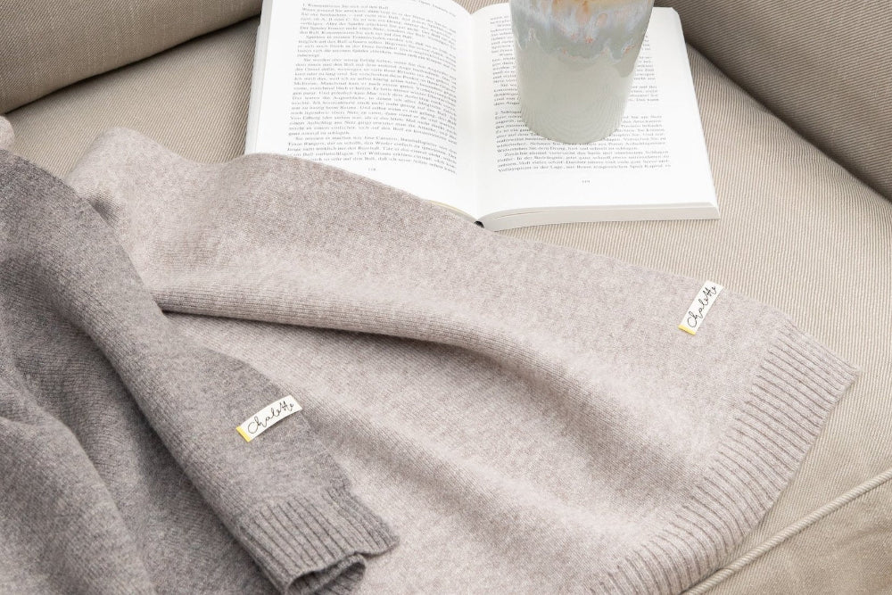 Eine graue und eine dunkelgraue Sofadecke liegen auf einem Sofa. Im Hintergrund ist ein Buch und eine Tasse