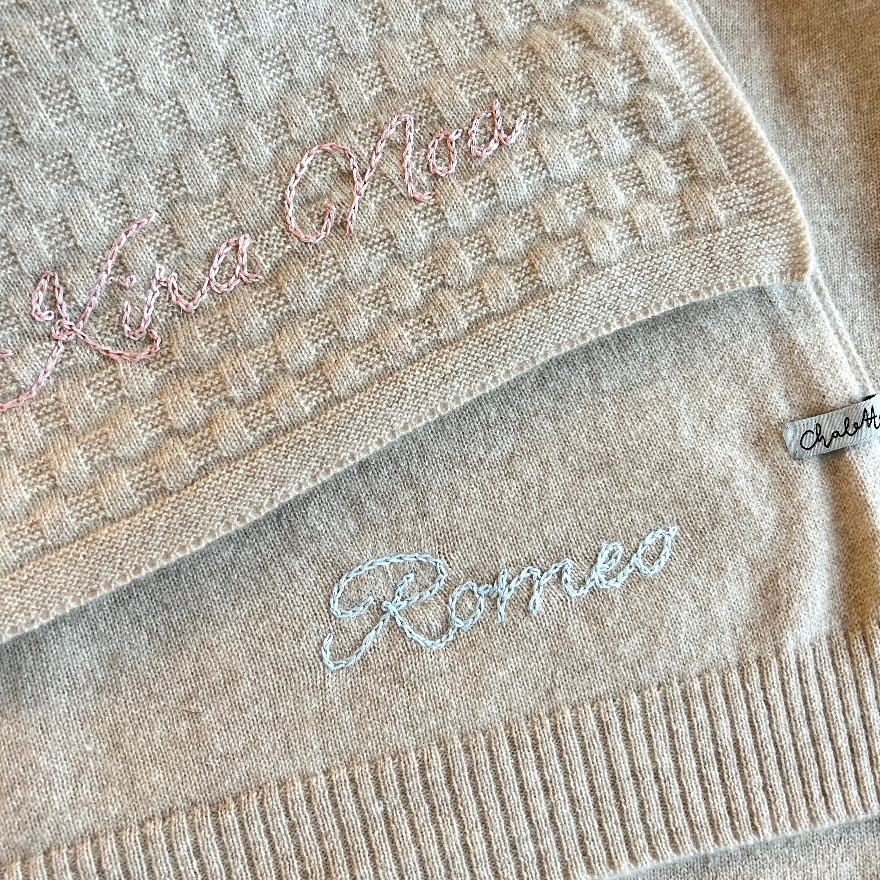 Eine beige Kaschmirdecke mit Waffelmuster mit handbestickten Namen "Kira Noa" in Rosa drapiert über einer beigen Kaschmirdecke mit handbesticktem Namen "Romeo" in Blau