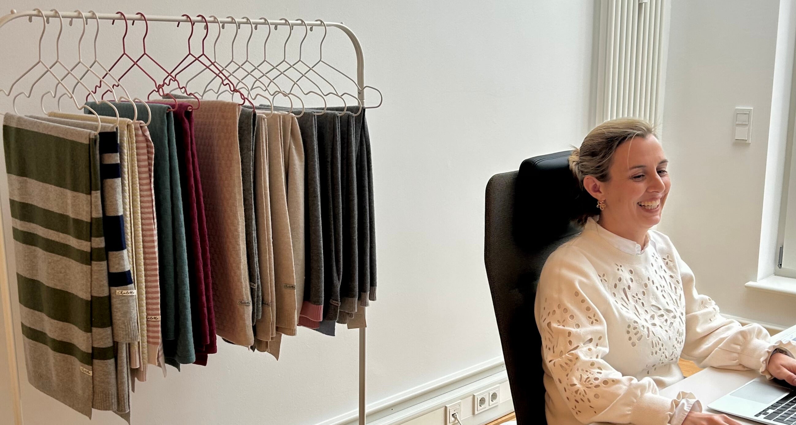 Geschäftsführerin von Chalette sitzt am Schreibtisch, Hintergrund mit Kaschmirdecken, die an Kleiderbügeln an Kleiderstange hängen