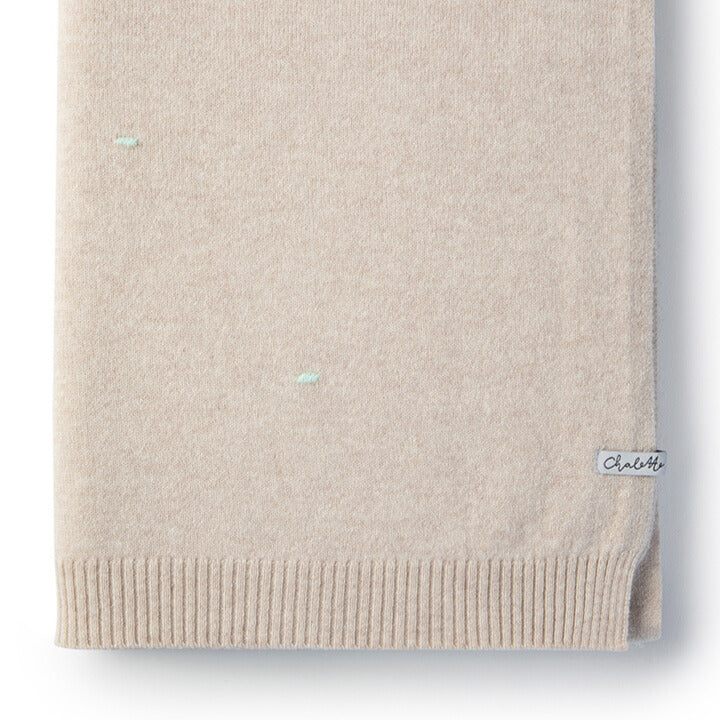 Detailaufnahme einer gefalteten beigen Babydecke mit türkisfarbenen Punkten mit sichtbarem Etikett: Chalette