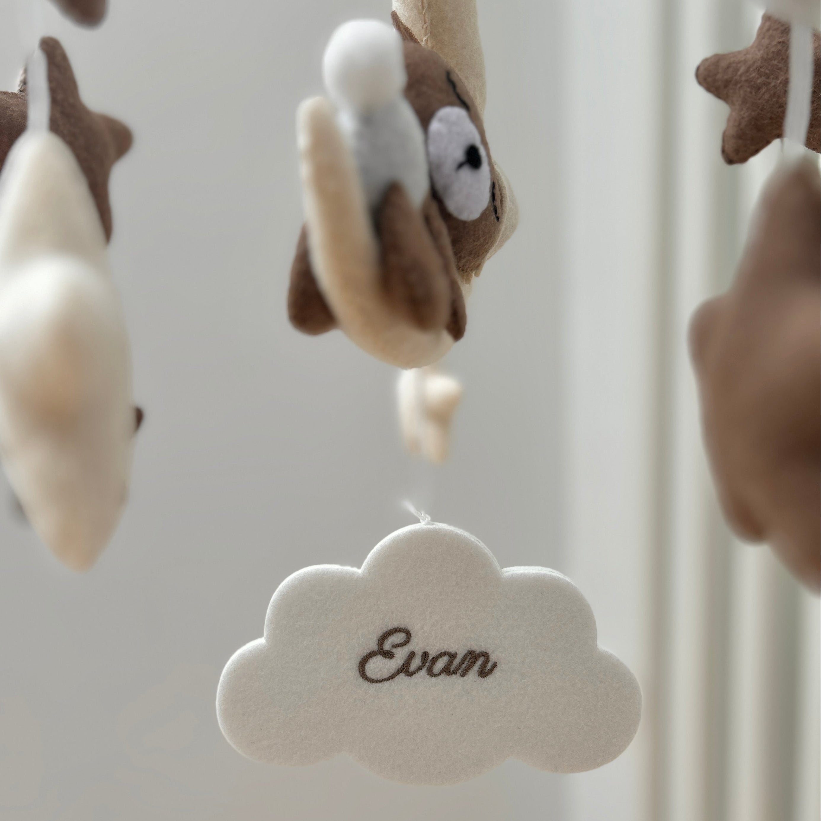 Mobile aus weichem Filz in warmen Beige- und Weiß-Tönen, in dessen Mitte eine helle Filzwolke hängt, bestickt mit den Namen „Evan“. Rundherum schweben kleine Filztiere, Sterne und Kugeln.