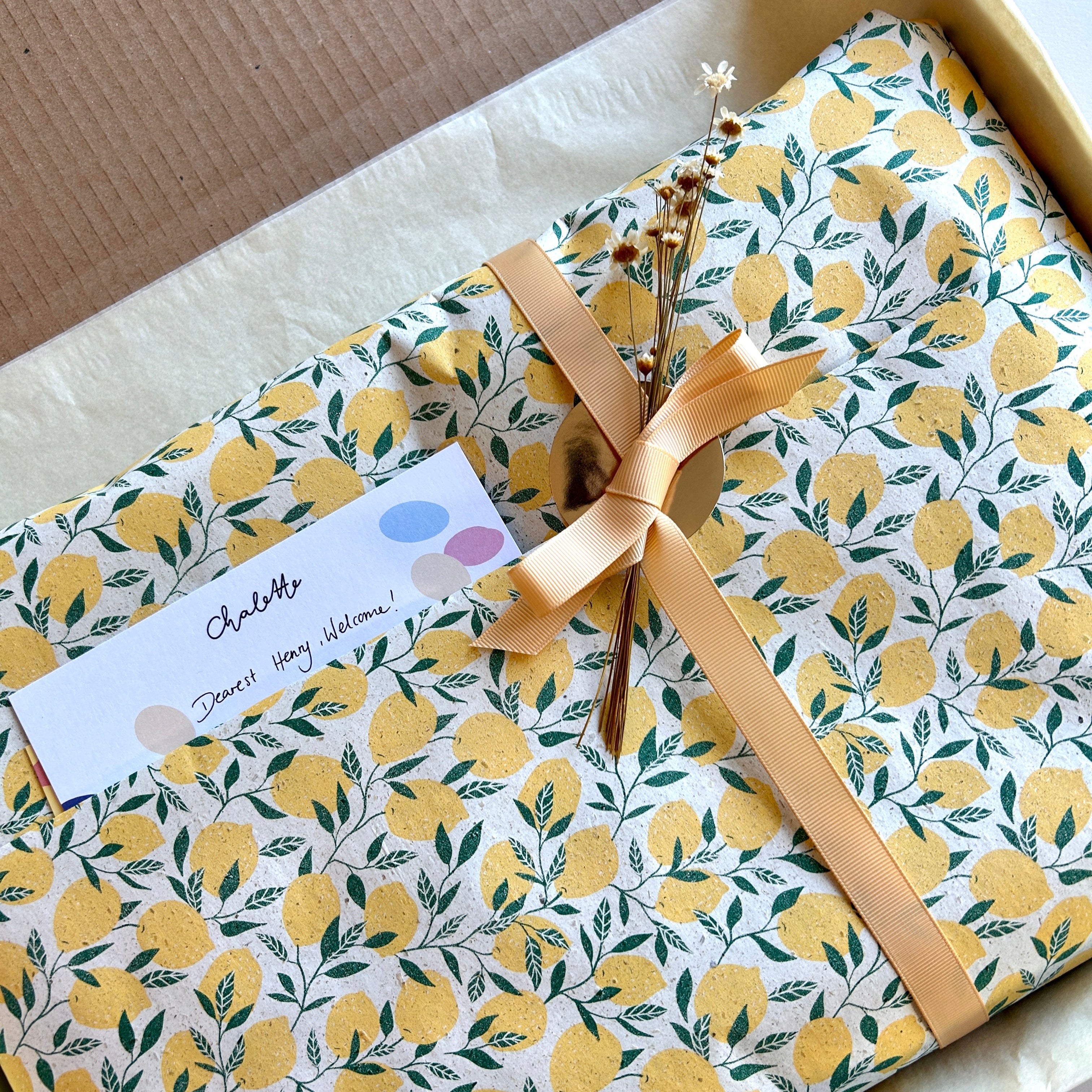 Geschenkverpackung mit Zitronenmusterpapier und beiger Schleife und weißen getrockneten Blumen, in offenem Karton liegend