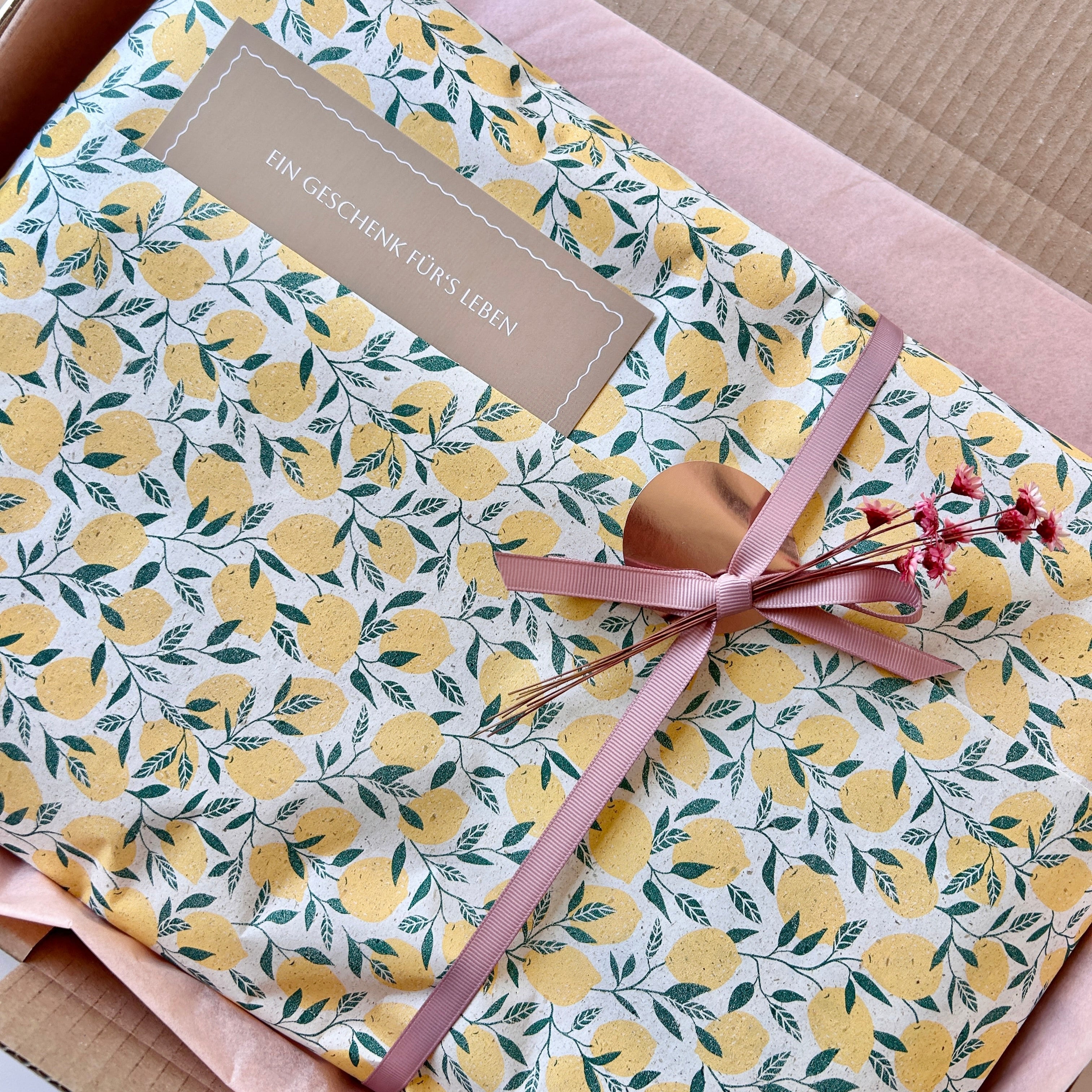 Geschenkverpackung mit Zitronenmusterpapier und rosafarbener Schleife, in offenem Karton liegend