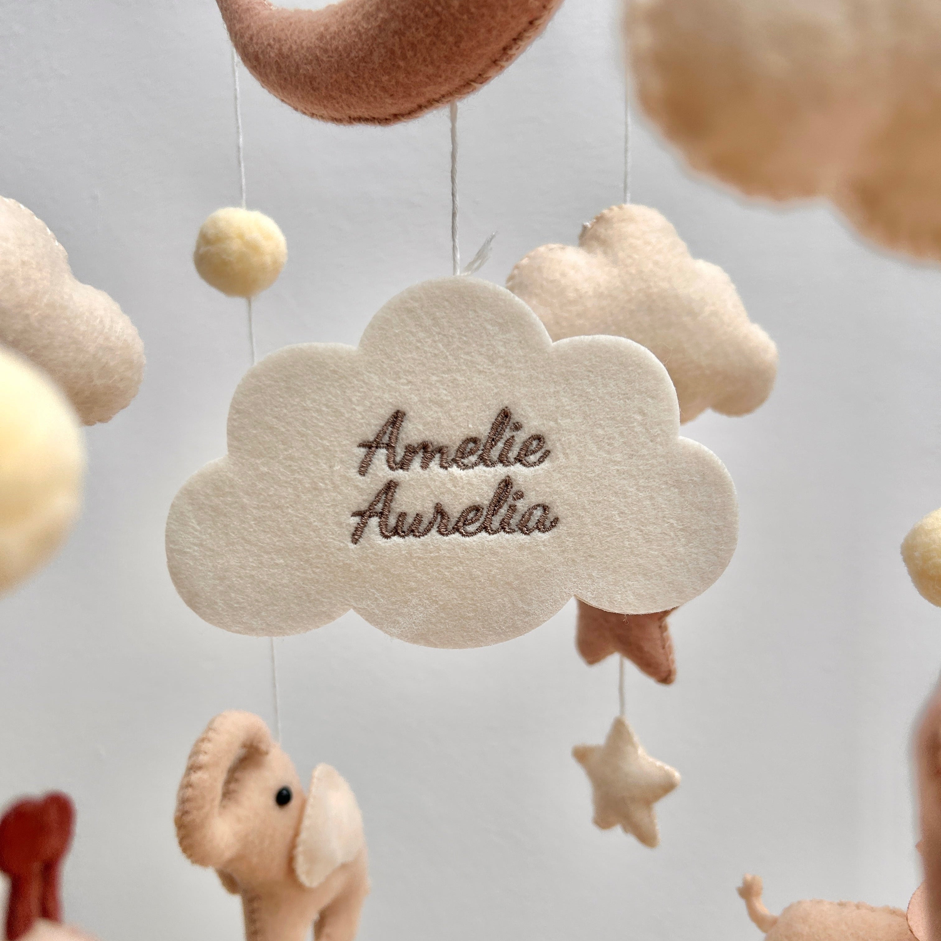 Mobile aus weichem Filz in warmen Beige- und Weiß-Tönen, in dessen Mitte eine helle Filzwolke hängt, bestickt mit den Namen „Amelie Aurelia“. Rundherum schweben kleine Filztiere, Sterne und Kugeln.