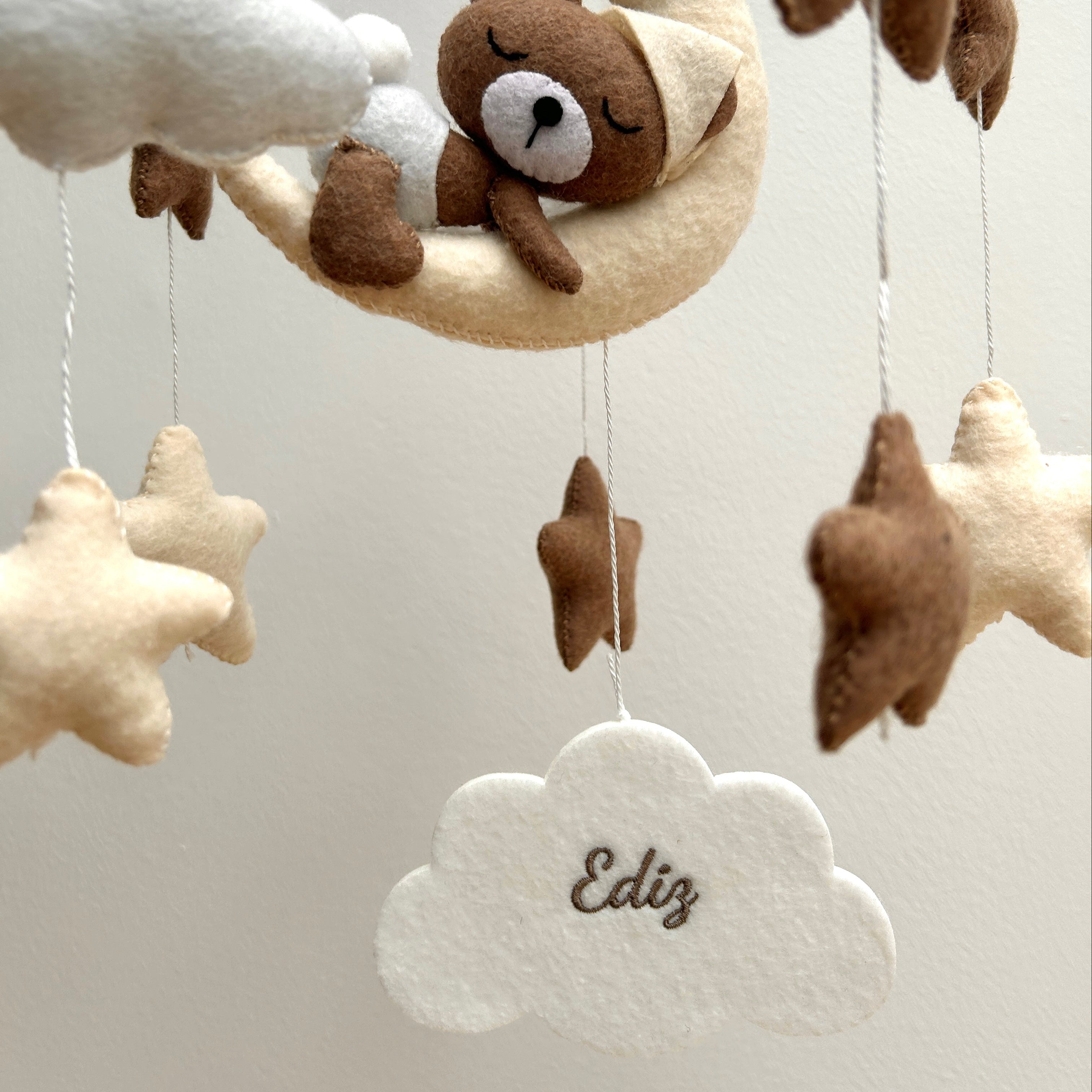 Mobile aus weichem Filz in Beige- und Weiß-Tönen, in dessen Mitte weiße Filzwolke hängt, bestickt mit den Namen "Ediz" in Braun. Darüber hängt ein Bär auf einer Wolke, rundherum hängen Sterne.