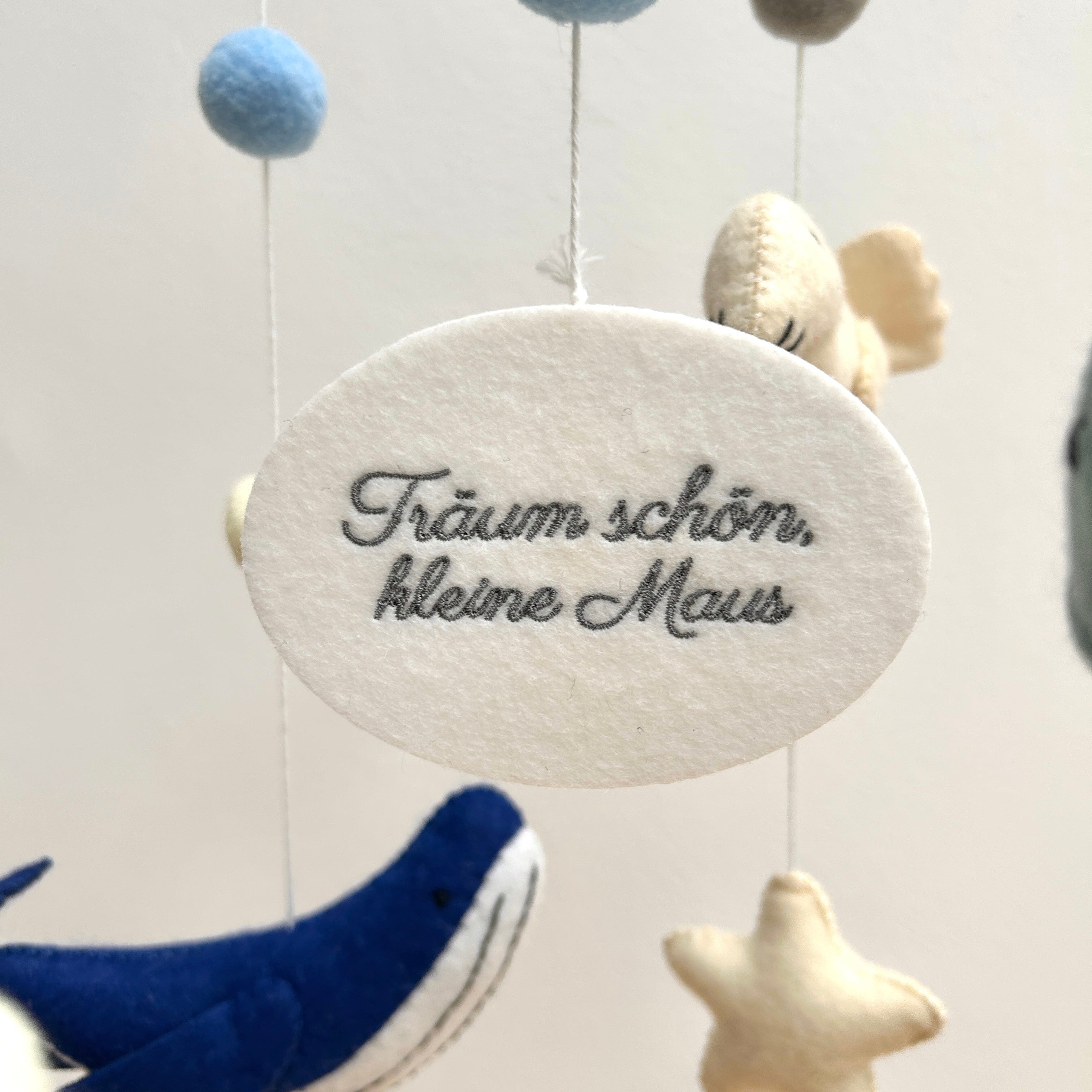 Mobile aus weichem Filz in Blau- und Weiß-Tönen, in dessen Mitte eine ovale Filzplatte in weiß hängt, bestickt mit dem Text "Träum schön, kleine Maus" in Grau. Rundherum hängen Meerestiere und Filzkugeln in Blau- und Weißtönen