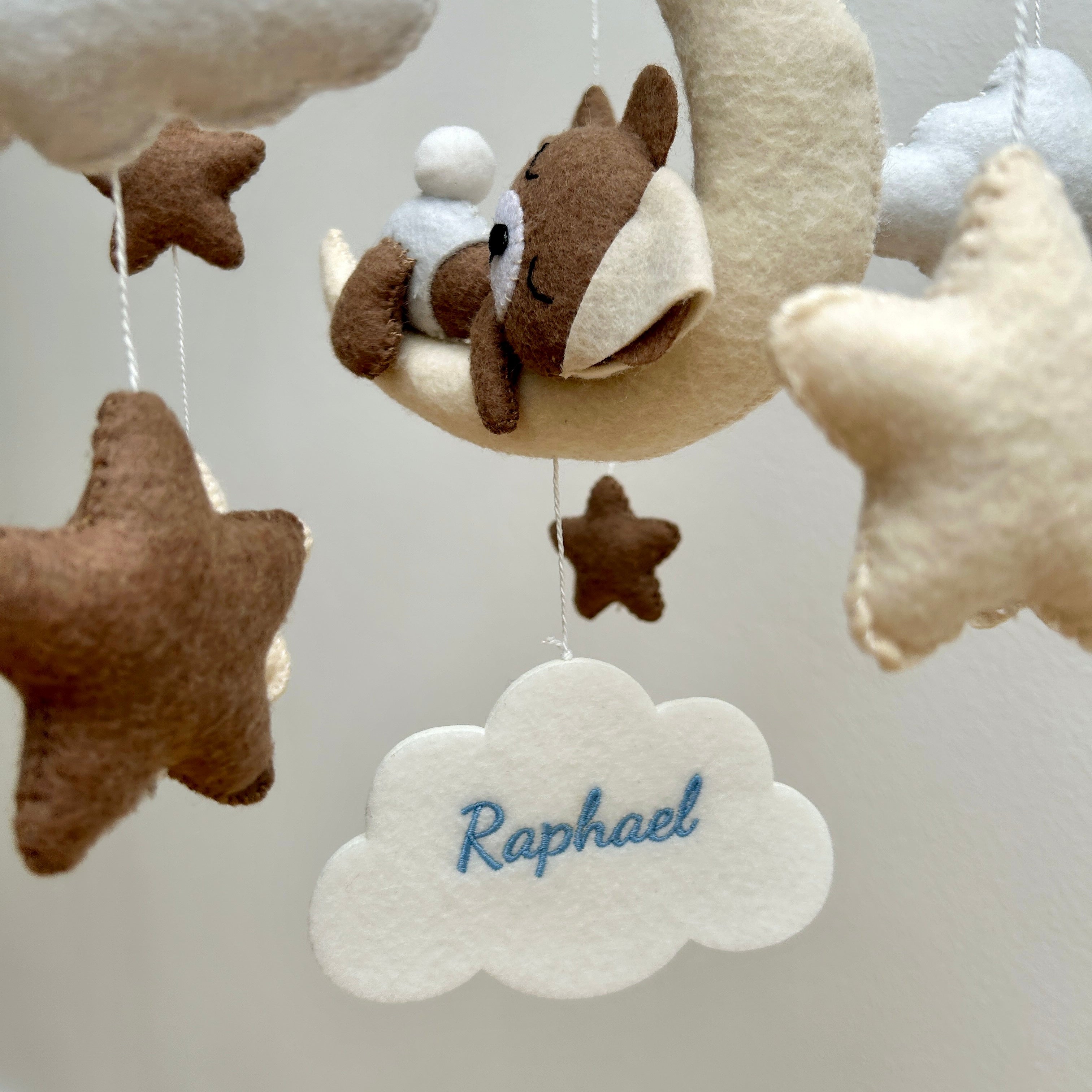 Mobile aus weichem Filz in Beige- und Weiß-Tönen, in dessen Mitte weiße Filzwolke hängt, bestickt mit den Namen "Raphael" in Blau. Darüber hängt ein Bär auf einer Wolke, rundherum hängen Sterne.