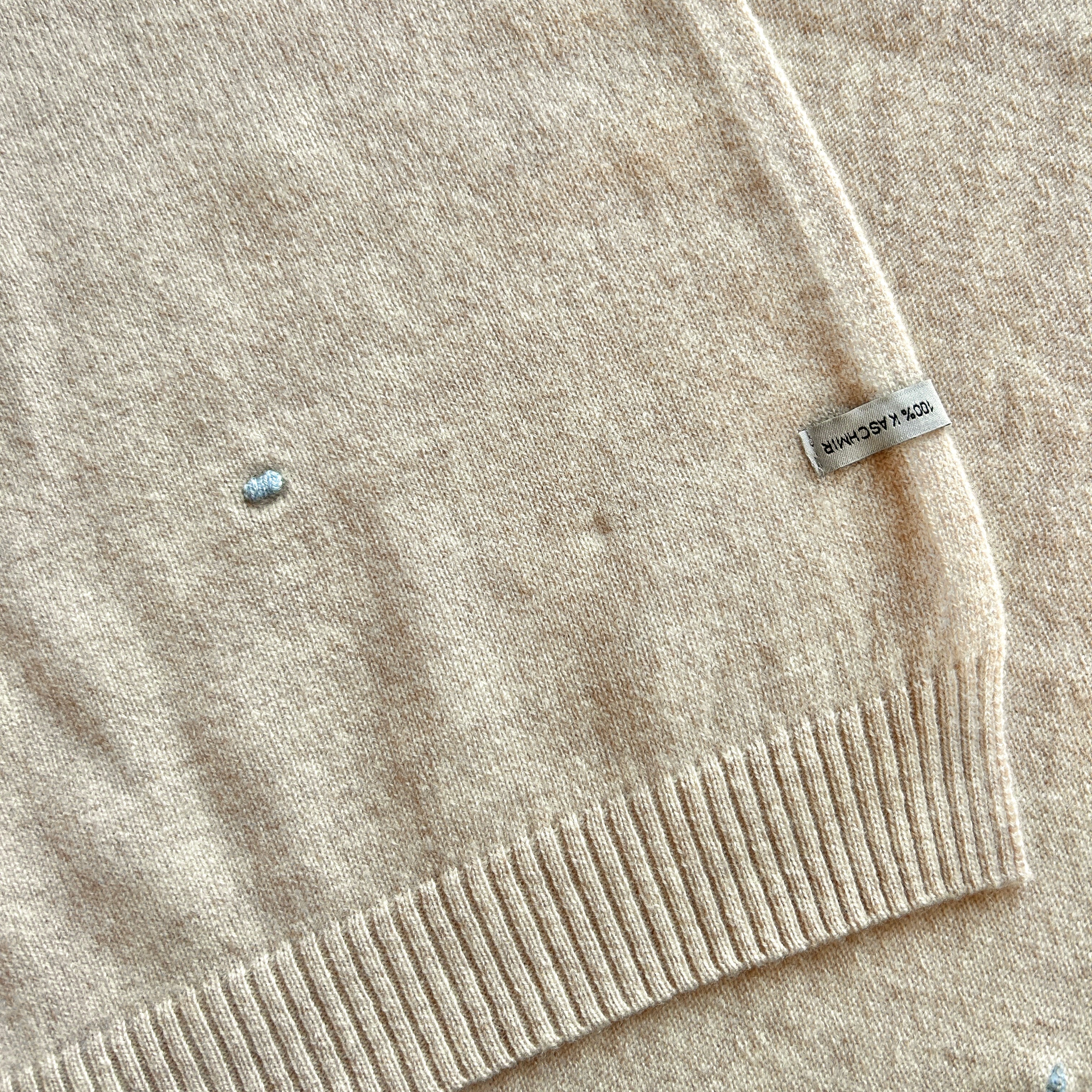 Ausschnitt einer Kaschmirdecke in Beige mit blauen Punkten. Das Etikett am Rand ist falsch aufgenäht.