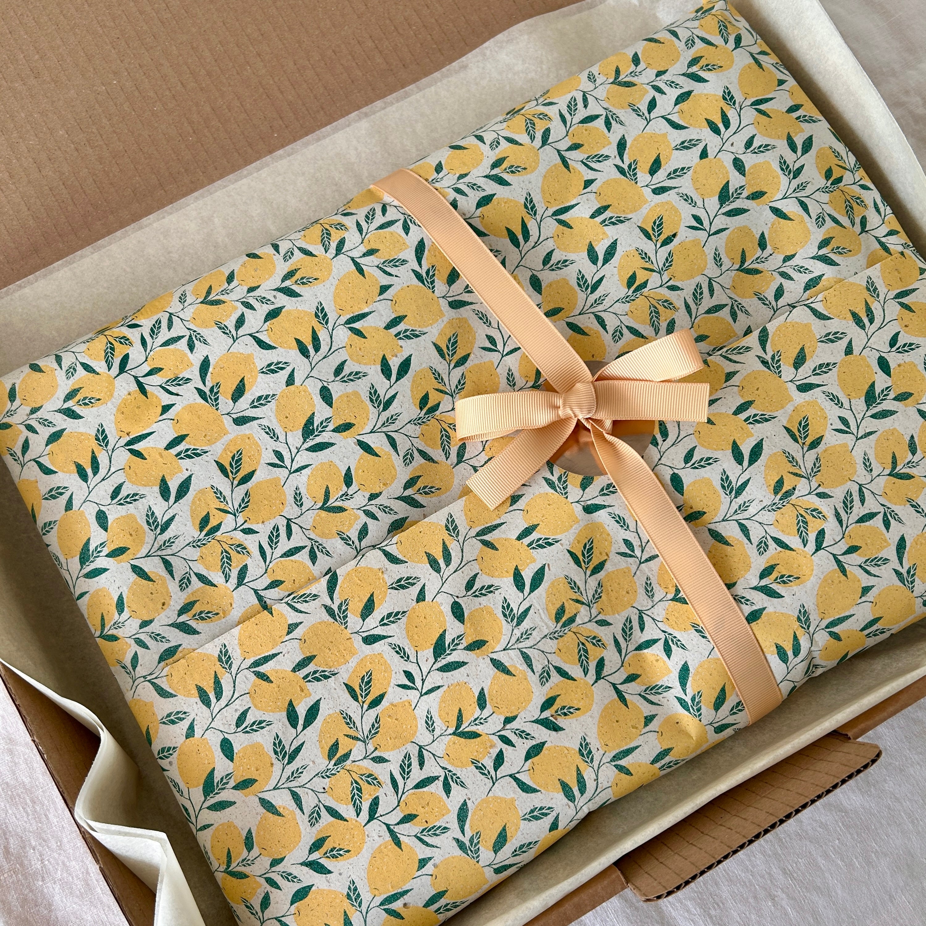 Geschenkverpackung mit Zitronenmusterpapier und beigefarbener Schleife, in offenem Versandkarton liegend