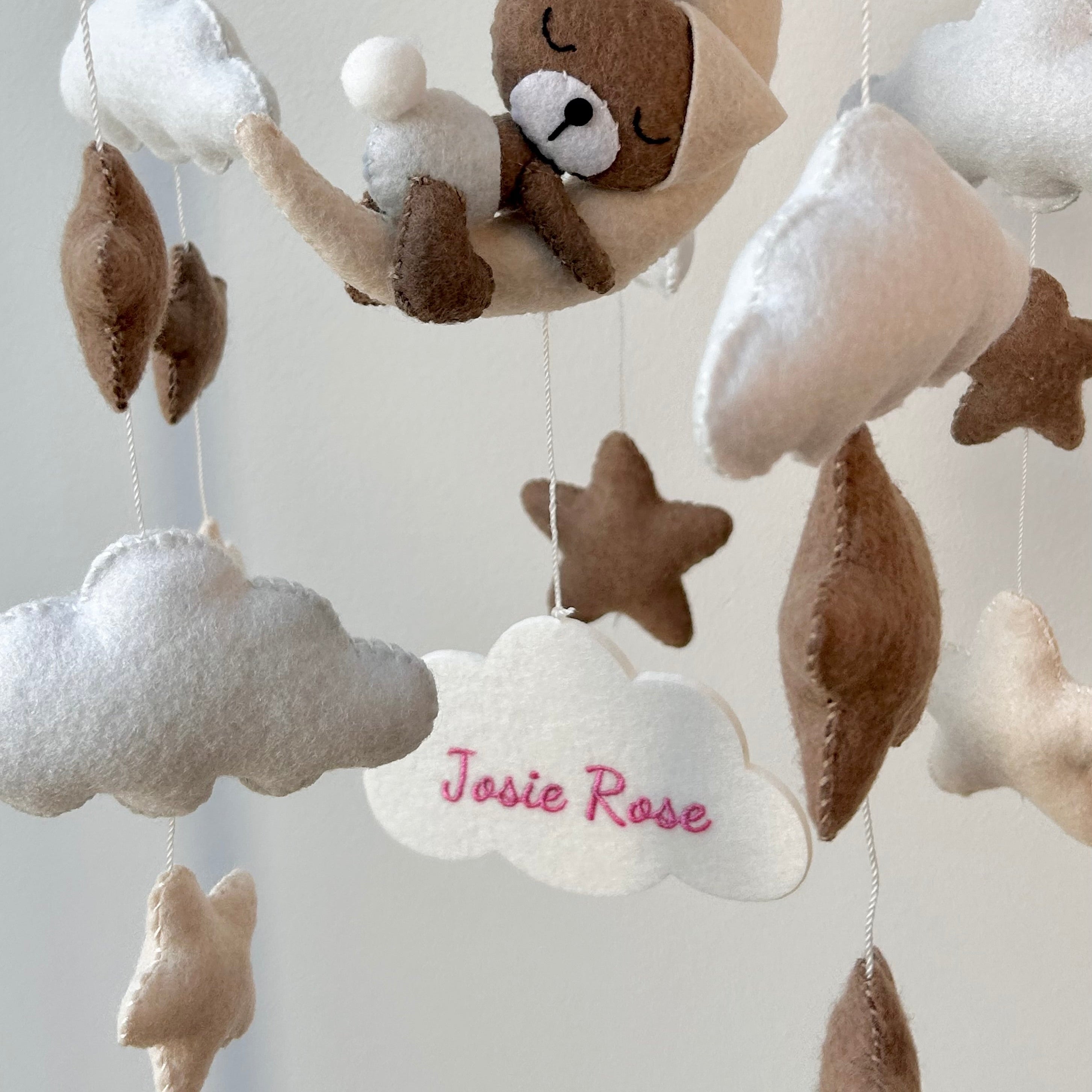 Mobile aus weichem Filz in warmen Beige- und Weiß-Tönen, in dessen Mitte eine helle Filzwolke hängt, bestickt mit den Namen „Josie Rose“. Rundherum schweben kleine Filztiere, Sterne und Kugeln.